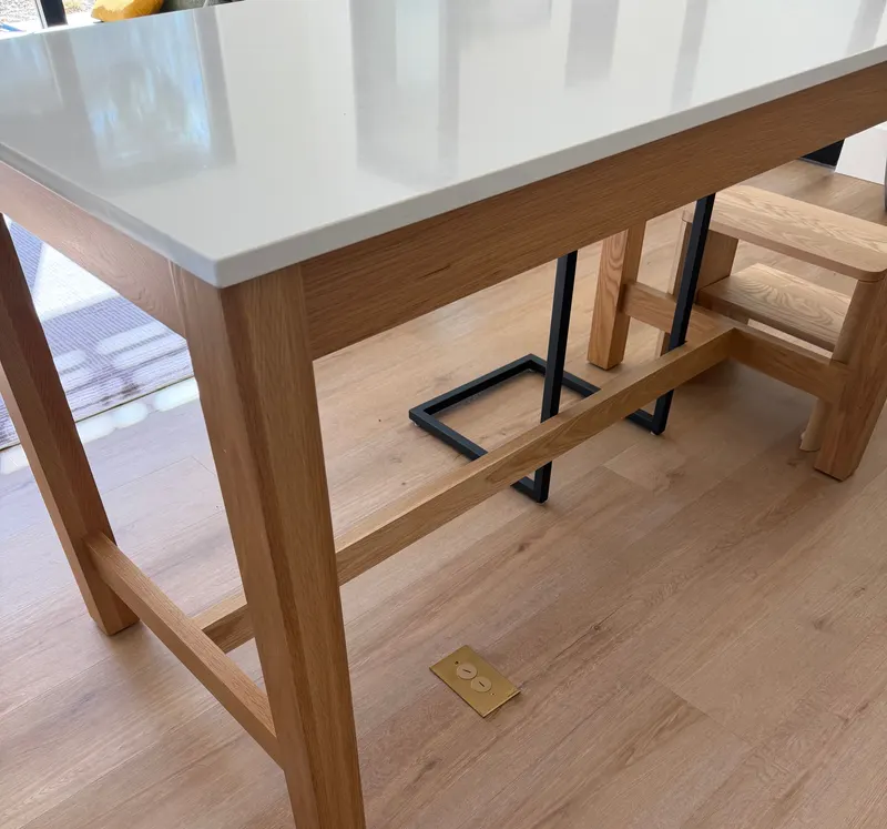 Brass floor outlet set flush into wood-look plank flooring beneath a movable oak island table with a white quartz top